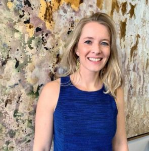 Caucasian blonde woman in a dark blue dress stands smiling in front of a earth-toned abstract painting