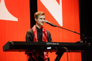 Caucasian male in black robes with a red sash performs at keyboard in front of a red screen with a white "N" in the background