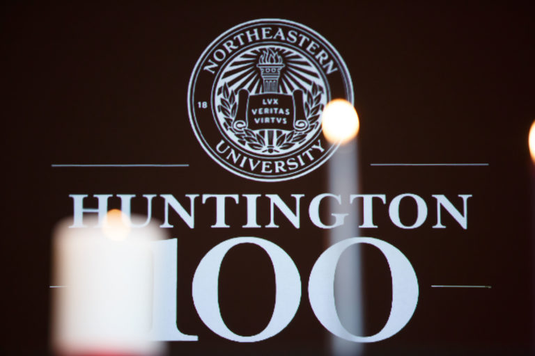 Two out of focus candles placed in front of the Northeastern seal and Huntinton 100 Logo
