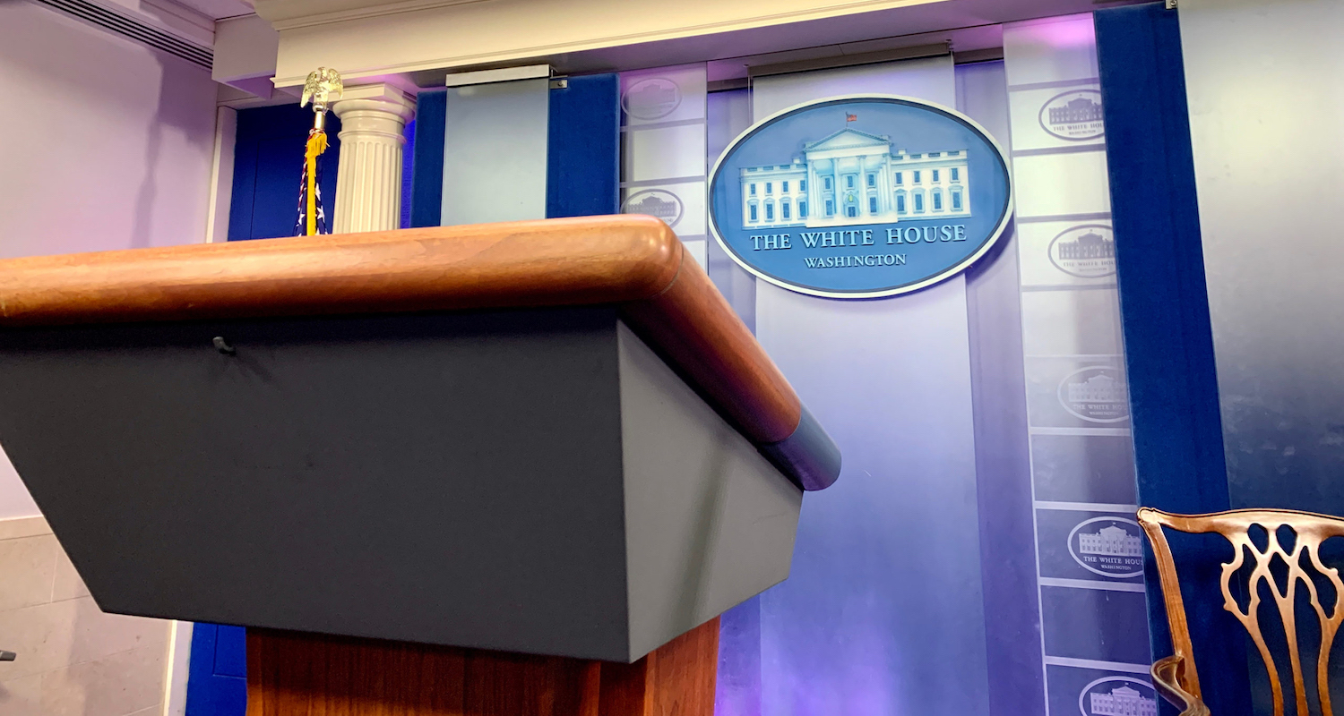 Podium in the foreground. Blue seal with White House emblem on the wall slightly out of focus in the background.