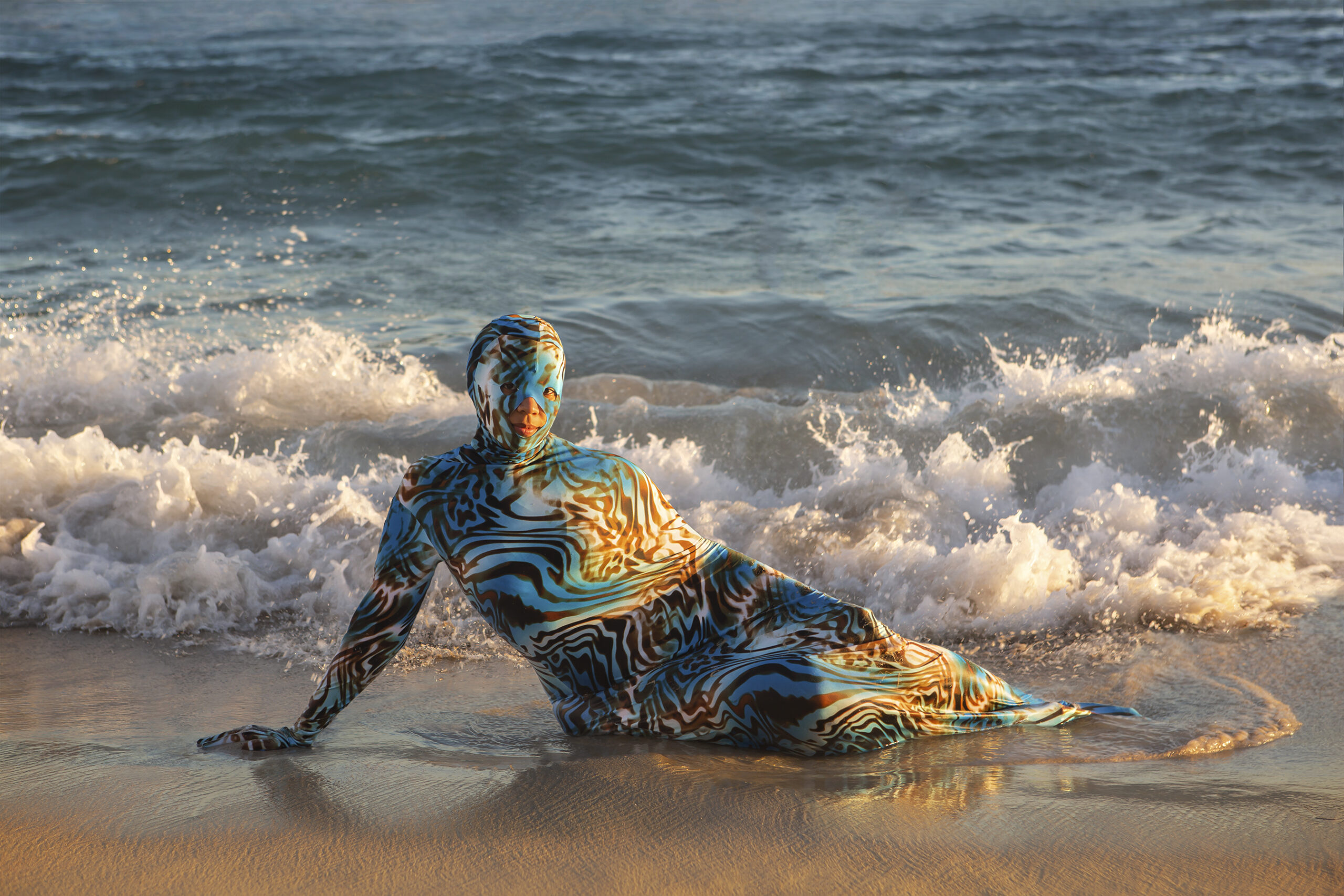 An image from Joiri Minaya's "Containers" photography series. Shows a woman lying on the beach, covered in a bodysuit.