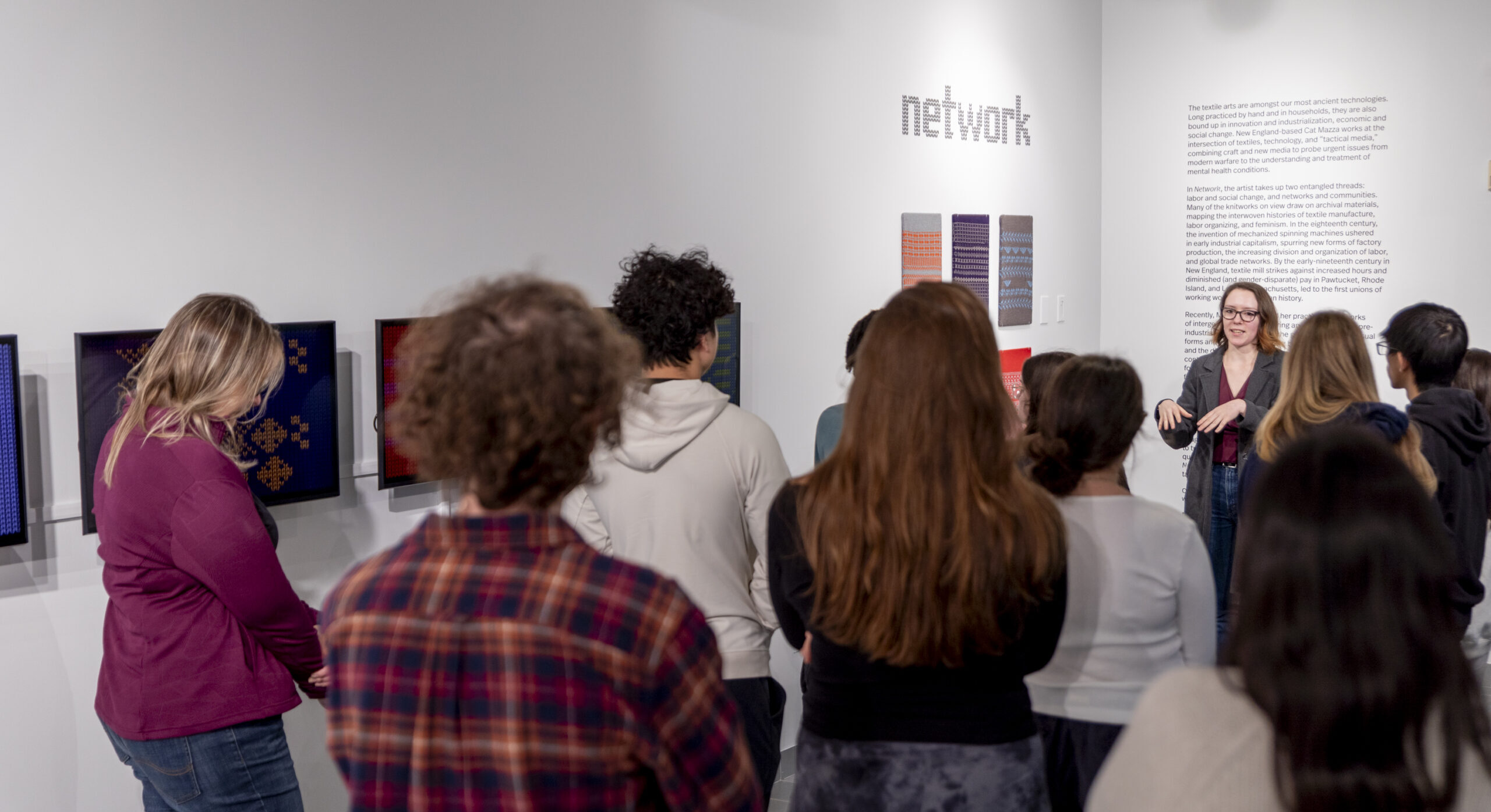 The image shows a group of people, mostly young adults, gathered in Gallery 360. They are examining and discussing the artwork on display, which includes colorful, abstract textile-like patterns. The central figure, a woman with short dark hair, is giving a presentation about the artwork. The text on the wall behind her reads "network," the title for Cat Mazza's 2024-2025 solo exhibition at Gallery 360.