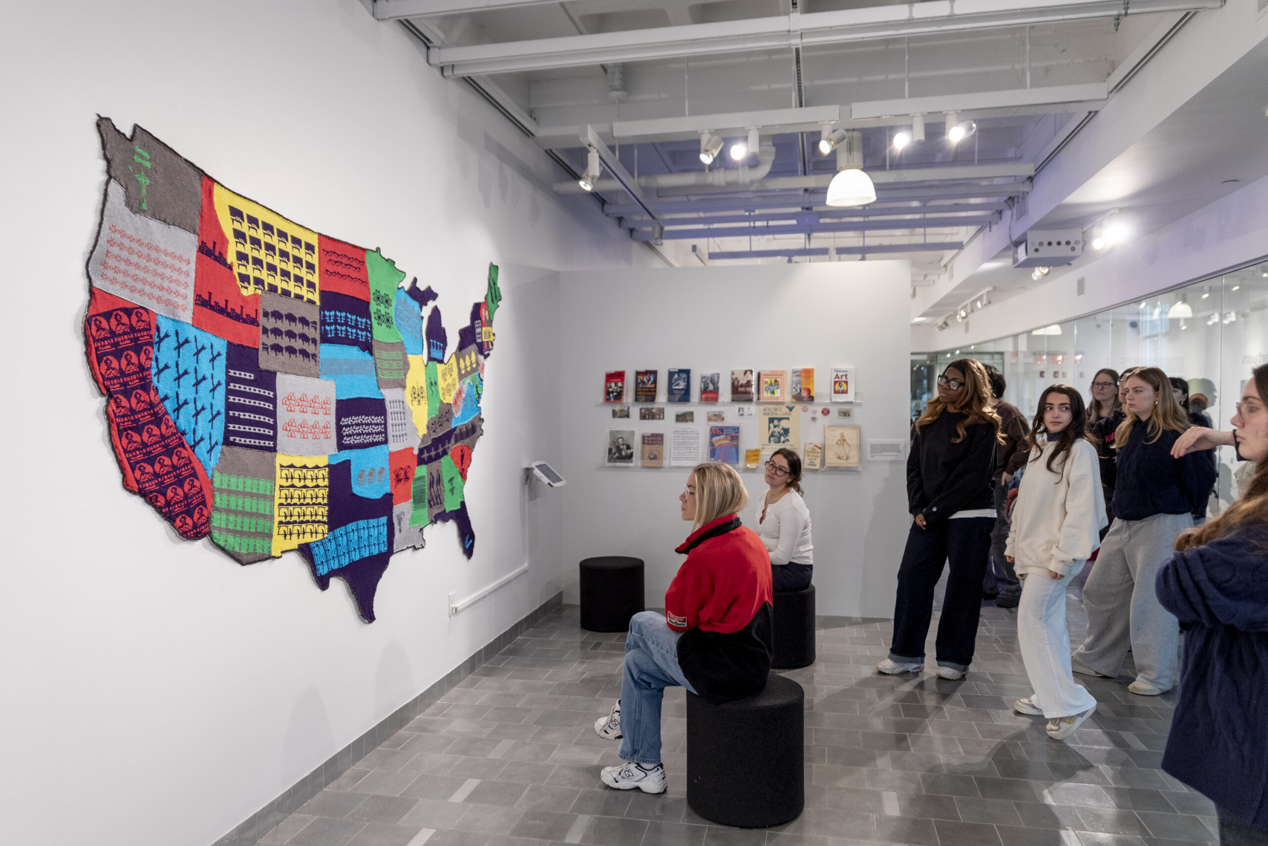 Photo shows Cat Mazza's massive textile art piece, the Labor Sister Sampler, in Gallery 360. To the right, a group of about 20 students looks at the piece.