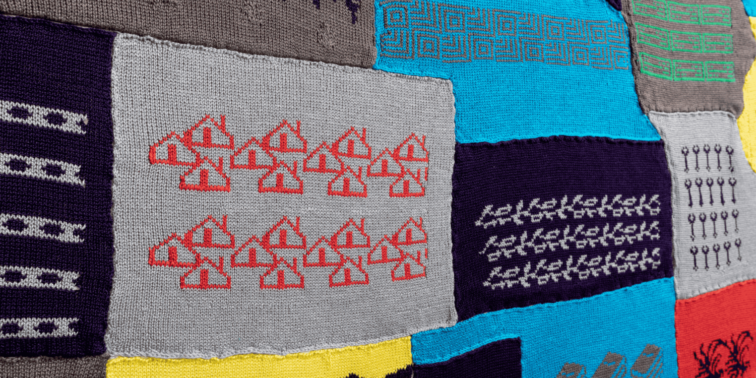 The image shows a close-up of "Labor Sister Sampler," a patchwork textile art piece by Cat Mazza. The material is made up of various patterns and designs, including geometric shapes, lines, and symbols in multiple colors such as gray, blue, yellow, and red. The intricate and colorful patterns create an aesthetically pleasing and visually interesting textile piece.