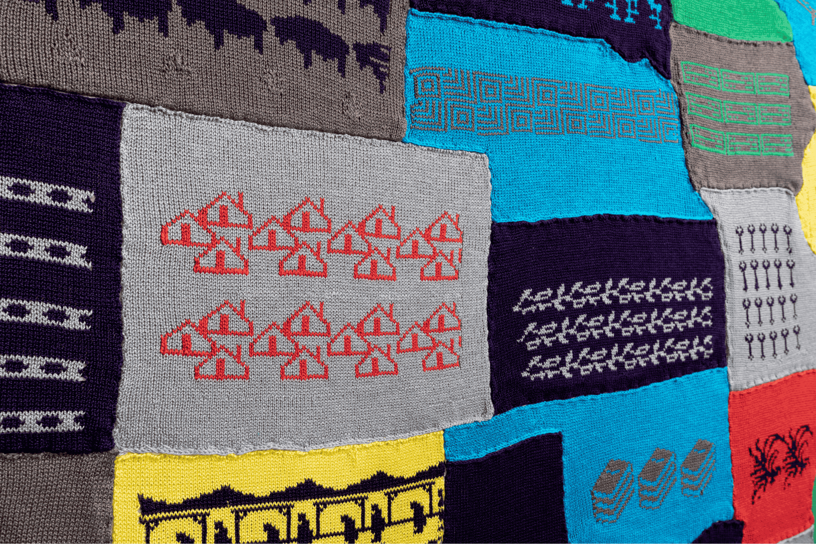 The image shows a close-up of "Labor Sister Sampler," a patchwork textile art piece by Cat Mazza. The material is made up of various patterns and designs, including geometric shapes, lines, and symbols in multiple colors such as gray, blue, yellow, and red. The intricate and colorful patterns create an aesthetically pleasing and visually interesting textile piece.